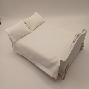 May include: A miniature white bed with two pillows. The bed frame has ornate detailing on the headboard and footboard. The bedding is a solid white color, creating a clean and simple aesthetic.
