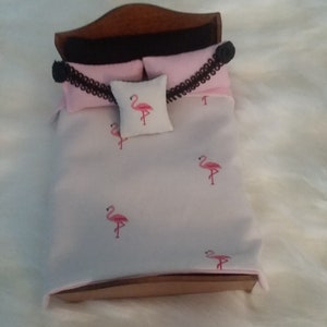 May include: A miniature doll bed with a white and pink flamingo patterned bedding set. The bed has a wooden headboard and footboard and a black and white patterned pillow.