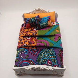 May include: A white miniature bed with a patterned quilt and pillows. The quilt is a patchwork of purple, green, orange, and blue fabrics with floral and paisley designs. The pillows are orange, purple, and blue with a patchwork design.