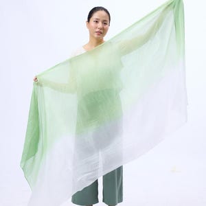 Könnte beinhalten: Ein hellgrüner und weißer Ombre-Schal aus einem durchsichtigen Stoff. Der Schal ist über die Schultern einer Person gelegt und wird in ihren Händen gehalten.