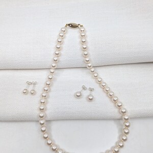 May include: A cream-coloured pearl necklace with a gold clasp and a small diamond, and two pairs of matching pearl earrings. The earrings are stud style. The pearls are a creamy white colour.