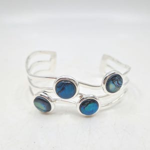 May include: A silver cuff bracelet with five round, bezel-set abalone shell cabochons. The abalone shells display iridescent blue and green hues. The bracelet has a three-band design and is displayed on a white surface.