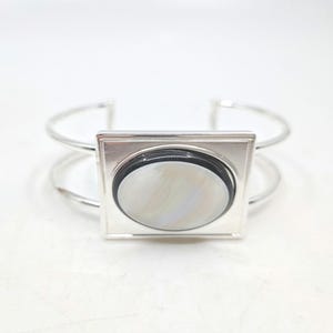 May include: A silver-toned cuff bracelet with a square centerpiece. The centerpiece features a black border around a pearlescent, oval-shaped inlay. The bracelet has a minimalist design, suitable for everyday wear.