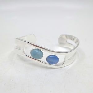 May include: A silver cuff bracelet featuring two oval, light blue gemstones. The bracelet has a modern, open design with a polished finish. The gemstones are set in a rectangular cutout in the center of the bracelet.