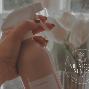 May include: A hand holding a light brown spray bottle with a white trigger and the text "ECO FRIENDLY" on the label. The bottle is in front of a white orchid plant and a round logo that says "MEADOW MAIDS HOUSE CLEANING".