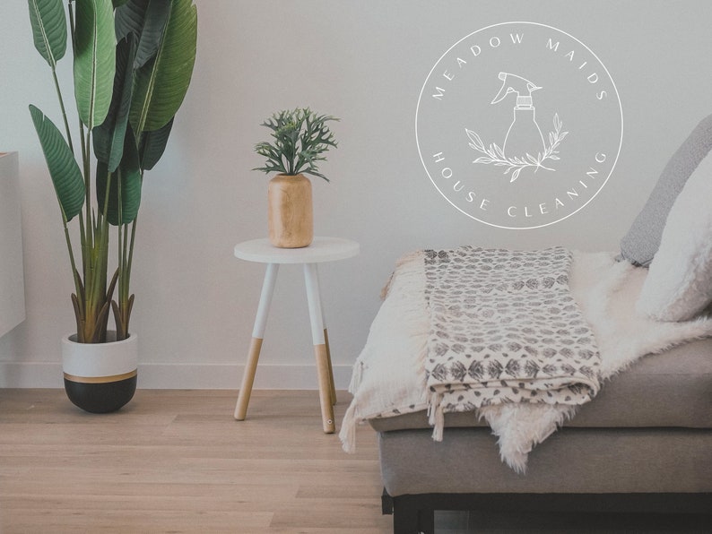 May include: A white circle logo with the text "Meadow Maids House Cleaning" and a line drawing of a spray bottle with leaves. The logo is on a white wall behind a grey sofa with a white throw blanket and a white side table with a green plant in a wooden pot.