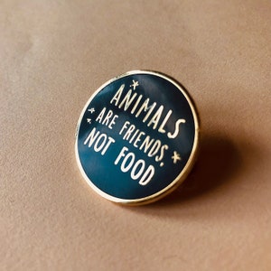 A round enamel pin on a plain background with the words animals are friends, not food written inside. There are 4 tiny stars surrounding the text. The background is black and the letters, outline and stars are gold colored and shiny.