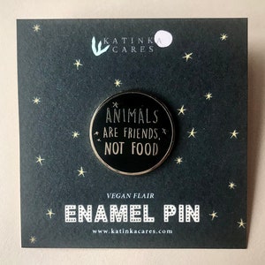 A round enamel pin attached to a black square backing card with small stars surrounding the pin. The pin has the words animals are friends, not food, some tiny stars and an outline in gold color. It has a black background in the circle.