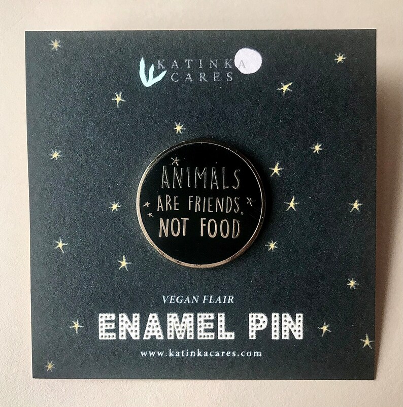 A round enamel pin attached to a black square backing card with small stars surrounding the pin. The pin has the words animals are friends, not food, some tiny stars and an outline in gold color. It has a black background in the circle.