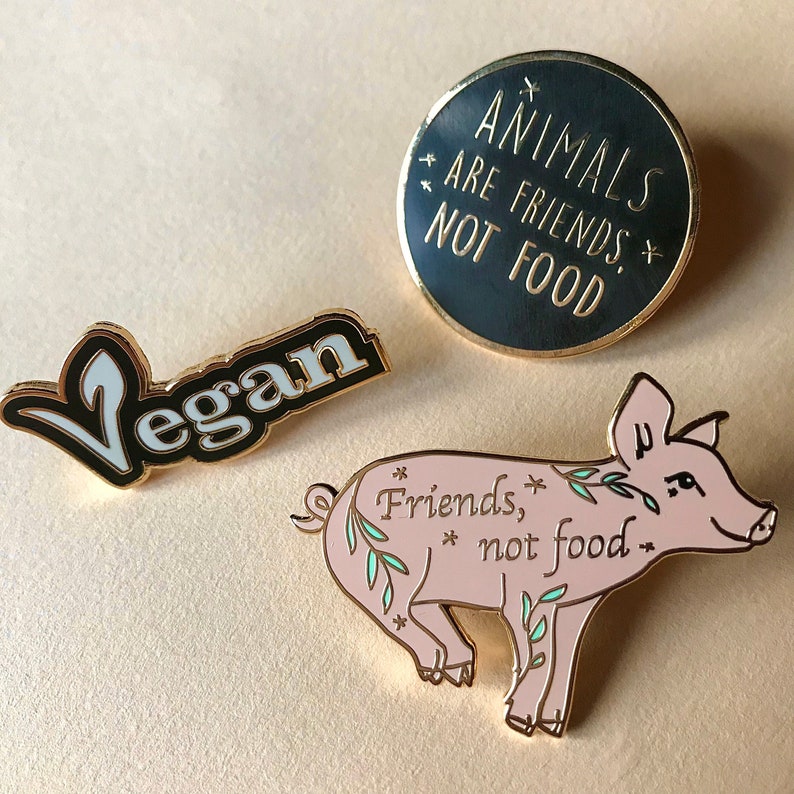 Picturing three different enamel pins. A black, white & gold colored pin that says vegan and the V made as the vegan logo, a black & gold round pin that says animals are friends not food and a cute pink pig pin with the words friends not food.