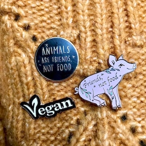 A close up of the three enamel pins attached to someone’s yellowish knitted sweater.