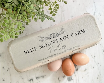 May include: A cardboard egg carton with the text "BLUE MOUNTAIN FARM" and "Fresh Eggs" printed on it. The carton is labeled "HAND GATHERED, ORGANIC, FREE RANGE". Three brown eggs are sitting in the carton.