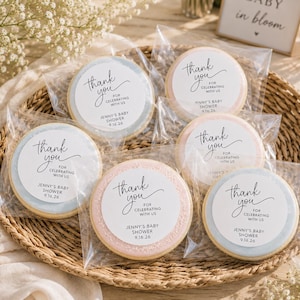 May include: A woven basket holds six round cookies individually wrapped in clear plastic. Each cookie has a white center with the words "thank you" and "for celebrating with us" in black script. The cookies are decorated with pastel blue and pink borders.