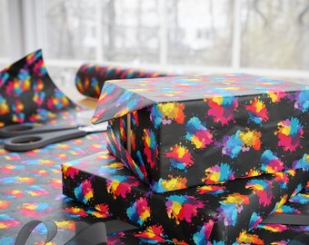 May include: A rectangular gift box wrapped in black paper featuring a vibrant multicolored paint splatter pattern in shades of blue, yellow, pink, and purple. The box sits on a matching sheet of wrapping paper with a black ribbon nearby.