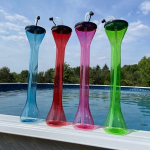 May include: Four tall, clear plastic cups with built-in straws. The cups are in different colors: blue, red, pink, and green. The cups are sitting on the edge of a swimming pool.