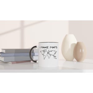 May include: White ceramic mug with a black handle and rim. The mug features a black and white world map design and the text "I MAKE MAPS". The mug is sitting on a white surface.