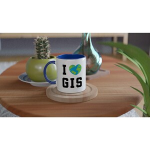 May include: White ceramic mug with a blue interior and handle. The mug features the text "I GIS" with a heart-shaped earth graphic. The mug is sitting on a wooden coaster on a wooden table. A cactus and a decorative vase are in the background.