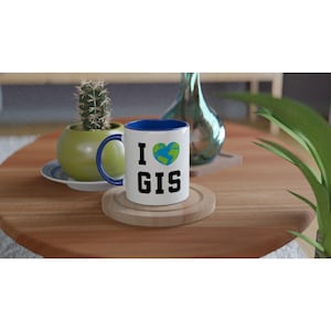 May include: White ceramic mug with a blue interior and handle, featuring the text "I GIS" with a heart-shaped Earth graphic. The mug sits on a wooden coaster, with a cactus and decorative vase in the background.