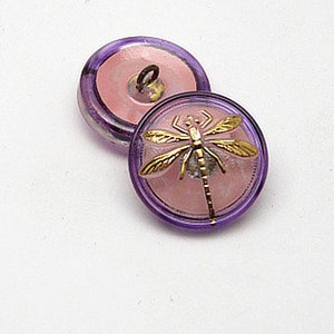 May include: Two round buttons with a dragonfly design. The buttons have a light pink center, a gold dragonfly, and a transparent purple rim. The buttons are on a white surface.