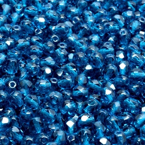 May include: A close-up view of a large quantity of small, faceted, teal blue glass beads. The beads are scattered randomly and create a shimmering effect.