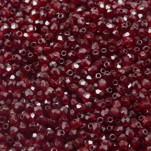 May include: A close-up view of a large quantity of small, faceted, red glass beads. The beads are arranged in a random pattern, creating a textured surface.