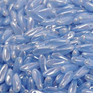 Puede incluir: Primer plano de un montón de cuentas de vidrio azul claro iridiscente. Las cuentas tienen forma de grano de arroz y un acabado liso y brillante.