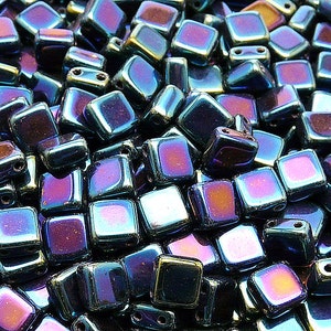 May include: A pile of iridescent square beads with a metallic finish. The beads have a mix of purple, blue, and gold hues, and are designed with two holes for stringing. These beads are suitable for jewelry making and crafting projects.