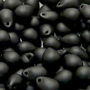 May include: A close-up of a pile of black glass beads in a teardrop shape. The beads have a matte finish and are scattered randomly.