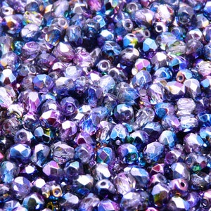 May include: A close-up image of a large quantity of iridescent blue and purple glass beads. The beads are faceted and have a shiny, reflective surface.