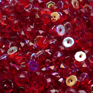 Op de afbeelding: Close-up van een verzameling gefacetteerde, dieprode kralen en iriserende pailletten. De kralen hebben meerdere facetten die het licht vangen, waardoor een sprankelend effect ontstaat. De pailletten zijn rond met een gat in het midden, wat textuur en dimensie toevoegt.