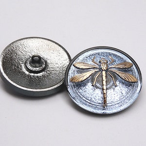 May include: A pair of silver-toned buttons with a dragonfly design. The dragonfly is gold and has a blue background. The button on the right is the front of the button and the button on the left is the back of the button.