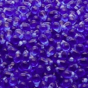 May include: A close-up image of a large quantity of small, teardrop-shaped glass beads. The beads are a vibrant, deep blue colour with a slight iridescent sheen.