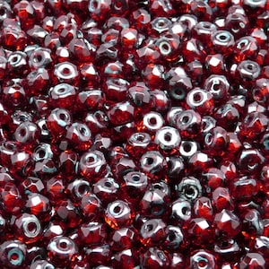 Może przedstawiać: Zbliżenie na stos małych, fasetowanych, czerwonych koralików szklanych. Koraliki mają delikatny, tęczowy połysk.