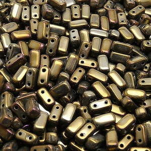 Puede incluir: Primer plano de un montón de cuentas rectangulares metálicas. Las cuentas son de color bronce dorado con destellos morados. Cada cuenta tiene dos agujeros para ensartar. Las cuentas están dispersas, creando una superficie texturizada.