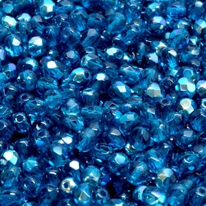 May include: A close-up image of a large quantity of small, faceted, iridescent blue glass beads. The beads are scattered randomly and have a reflective, shimmering quality.