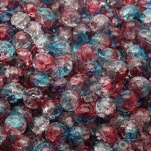 May include: A close-up of a pile of small, round, glass beads. The beads are a mix of clear, blue, and pink colors. The beads have a speckled, textured surface.