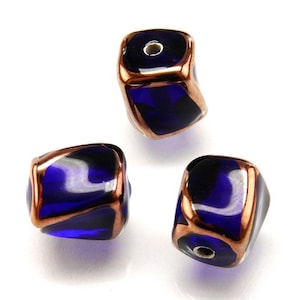 Puede incluir: Tres cuentas de vidrio azul oscuro con ribete de cobre. Las cuentas son de forma cuadrada con bordes redondeados.