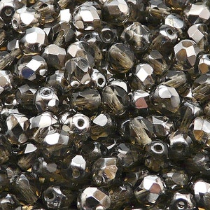 Puede incluir: Una vista de cerca de un montón de cuentas de vidrio facetadas de color gris oscuro. Las cuentas son pequeñas y tienen un acabado brillante y metálico.