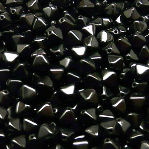 Puede incluir: Primer plano de un montón de cuentas negras facetadas. Las cuentas tienen forma triangular con múltiples lados y un acabado brillante. Cada cuenta tiene un pequeño agujero para ensartar. Las cuentas están densamente empaquetadas, llenando el encuadre.