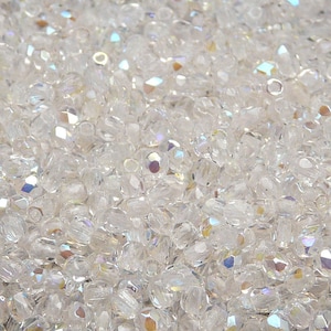 May include: A close-up image of a large quantity of clear glass beads with an iridescent finish. The beads are small and round, and they are scattered randomly across the surface.