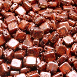 May include: A close-up of a pile of small, square, copper-colored glass beads. The beads have a metallic sheen and are slightly iridescent.