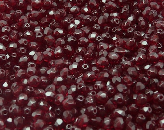 May include: A close-up view of a large quantity of small, faceted, dark red glass beads. The beads are arranged in a random pattern, creating a textured surface.