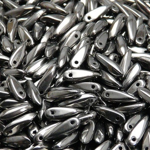 May include: A close-up shot of a pile of shiny, metallic teardrop-shaped beads. Each bead has a small hole at the top. The beads are a silver color and reflect light, creating a glossy appearance. The beads are scattered, filling the frame.