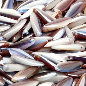 Könnte beinhalten: Eine Sammlung von irisierenden, perlmuttartigen und opaleszierenden Glasperlen in Braun-, Weiß- und Blautönen. Die Perlen sind reisförmig und haben eine glatte, polierte Oberfläche.