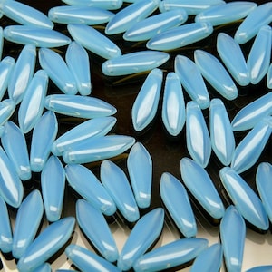 Puede incluir: Un gran grupo de cuentas de vidrio azul claro en forma de lágrima. Las cuentas están esparcidas sobre una superficie negra.