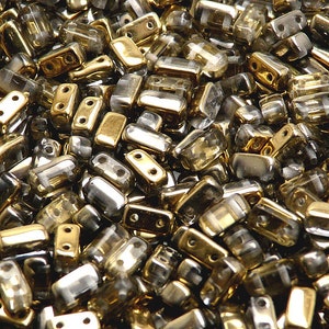 Peut inclure: Gros plan sur un tas de petites perles de verre rectangulaires. Les perles sont un mélange de couleurs claires et dorées, certaines ayant une finition métallique.