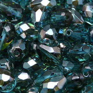 Puede incluir: Primer plano de cuentas de vidrio azul verdoso con un diseño facetado. Las cuentas son iridiscentes y tienen una variedad de formas y tamaños.