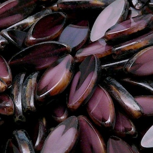 Può includere: Primo piano di un mucchio di perline di vetro viola e marrone. Le perline sono di forma ovale e hanno una finitura lucida.