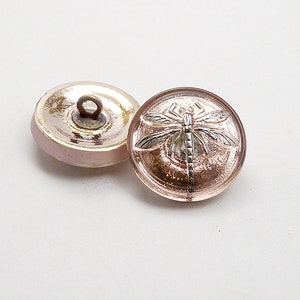 Puede incluir: Dos botones redondos de vidrio rosa con un diseño de libélula plateada. El botón de la derecha tiene un diseño de libélula en relieve. El botón de la izquierda tiene un vástago en la parte posterior.