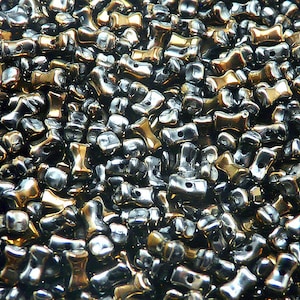 Może przedstawiać: Zbliżenie na stos metalicznych koralików. Koraliki są mieszanką kolorów srebrnego i złotego, o unikalnym, wydłużonym kształcie. Każdy koralik ma mały otwór, co sugeruje, że są przeznaczone do nawlekania. Koraliki mają błyszczącą, odblaskową powierzchnię.